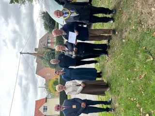 A group of eight people, two wearing chains of office, stand on a grass verge in Aston Abbotts holding the Gurney Cup and a certificate