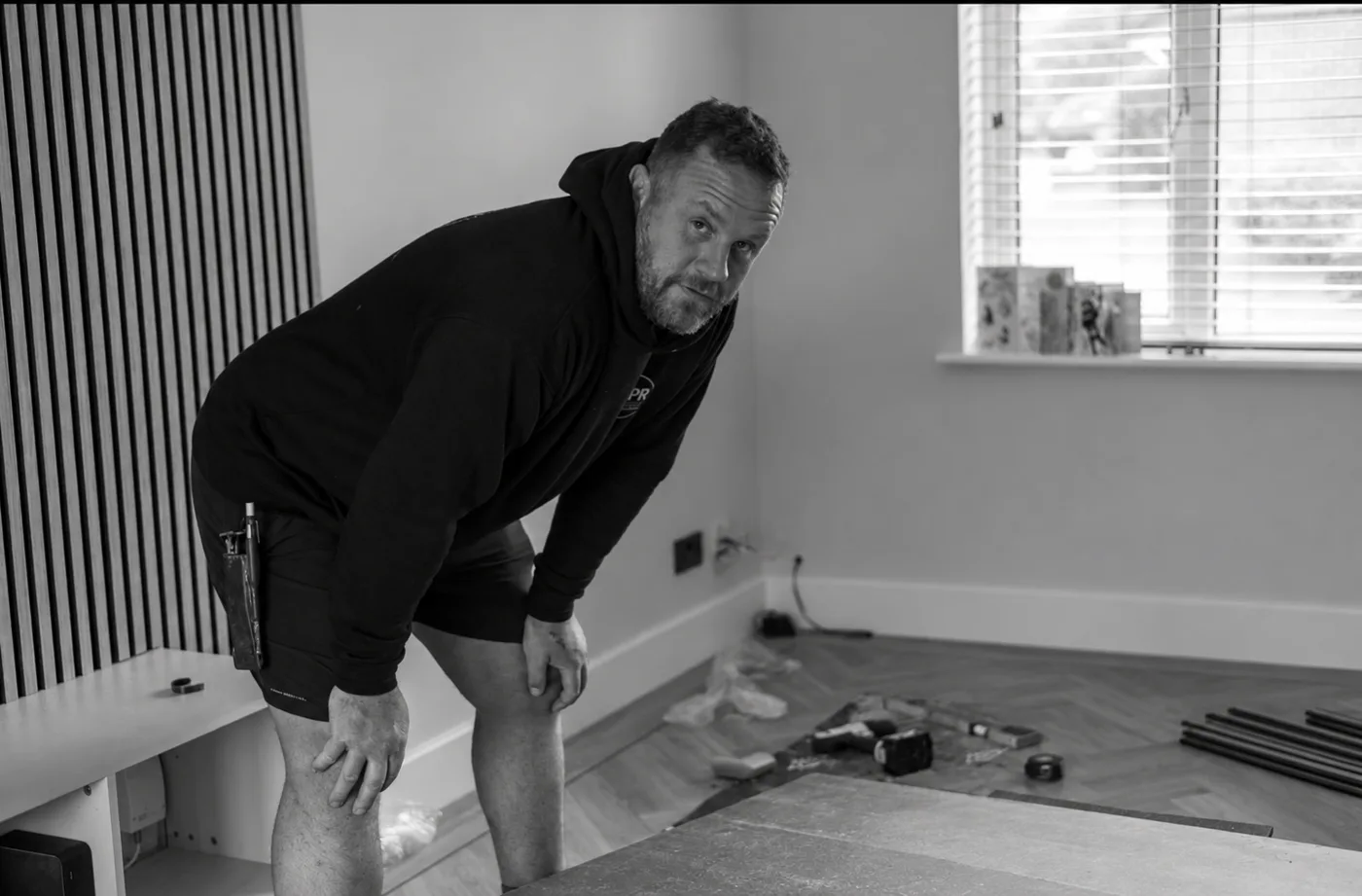 Black and white photograph of Shane Williams leaning on his knees in a room being renovated, with herringbone flooring, tools and a slatted wall panel around him
